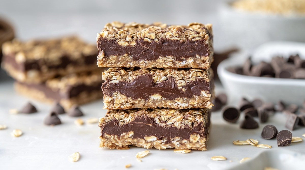 3 barritas de avena y chocolate apiladas una encima de otra, con algunos ingredientes como chocolate y copos de avena desenfocados al fondo.