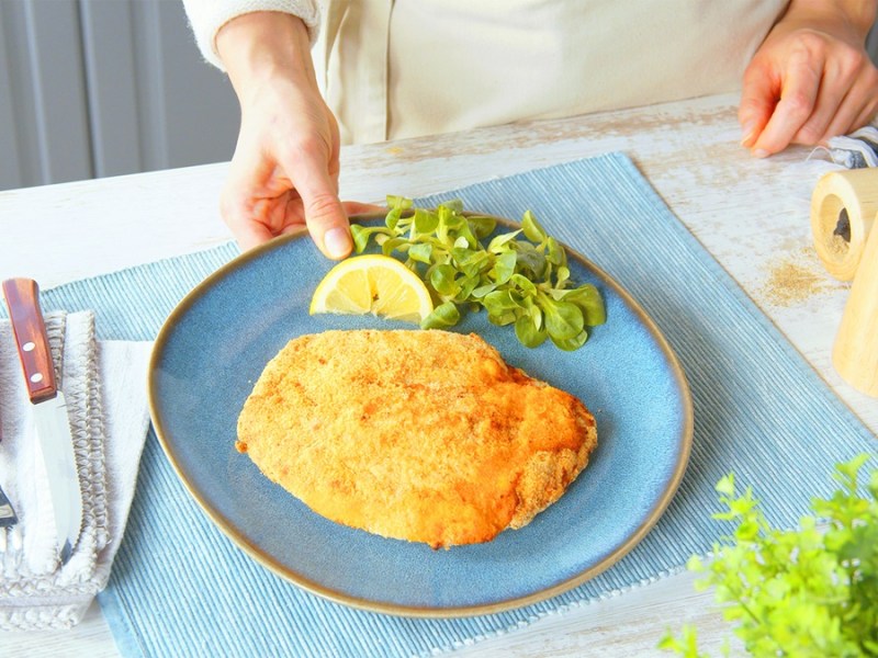 Un cordon bleu con salami en un plato azul. En el plato hay una rodaja de limón y un poco de canónigos. Junto al plato hay cubiertos y un salero y pimentero.