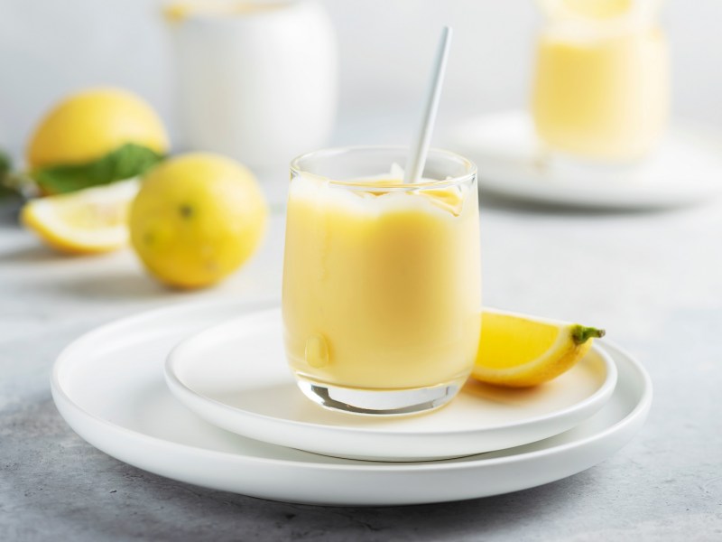 Un vaso de crema de limoncello en un plato, también una rodaja de limón, al fondo desenfocados dos vasos más y limones.