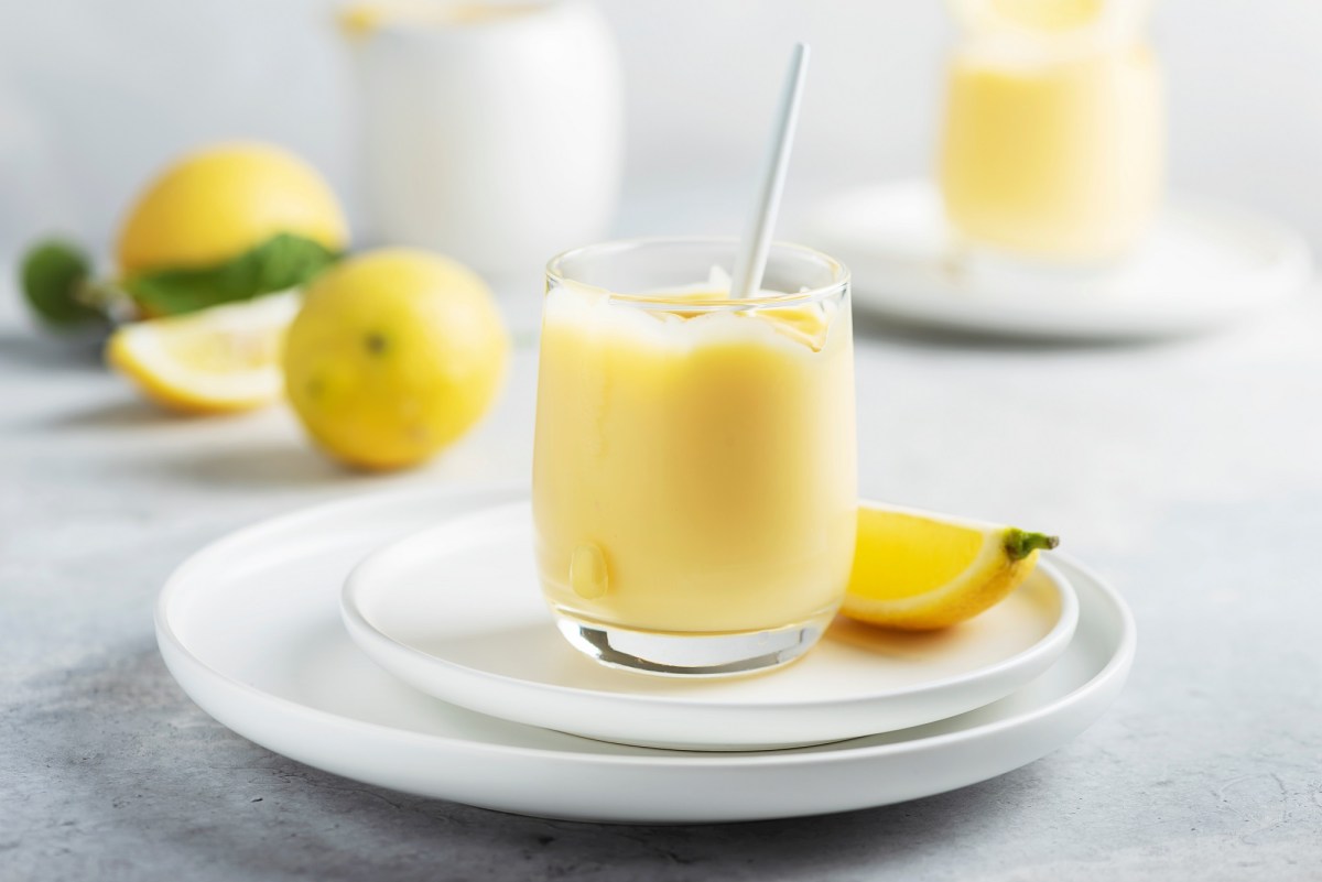 Un vaso de crema de limoncello en un plato, también una rodaja de limón, al fondo desenfocados dos vasos más y limones.
