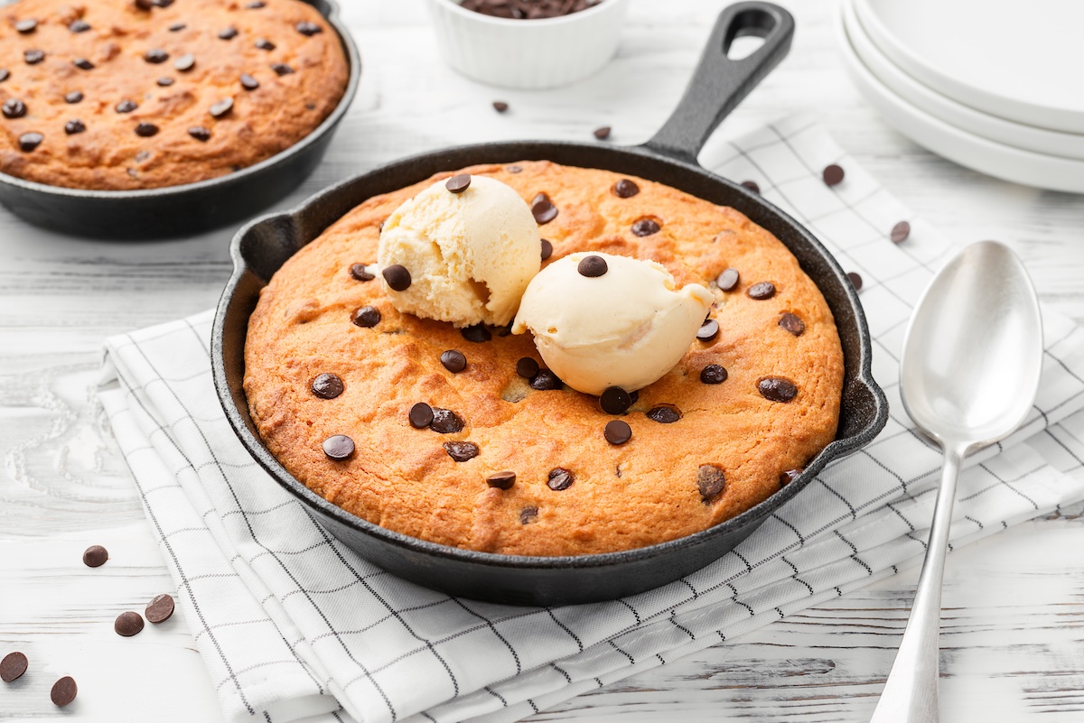 Una skillet cookie, es decir, una galleta en sartén, con chispas de chocolate en una sartén, decorada con dos bolas de helado. Junto a ella, una cuchara y otra sartén con una galleta.