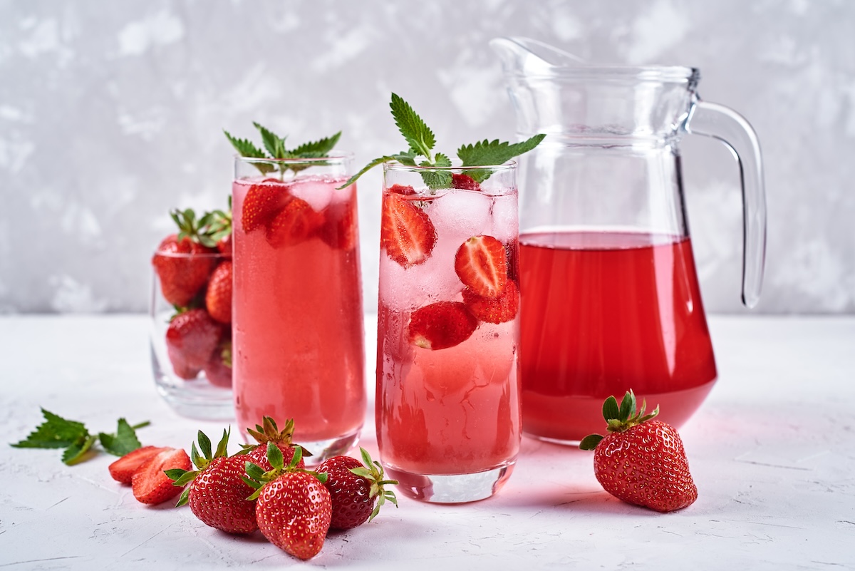 2 vasos de limonada de fresa con mango y plátano, junto a una jarra con la limonada y algunas fresas frescas.