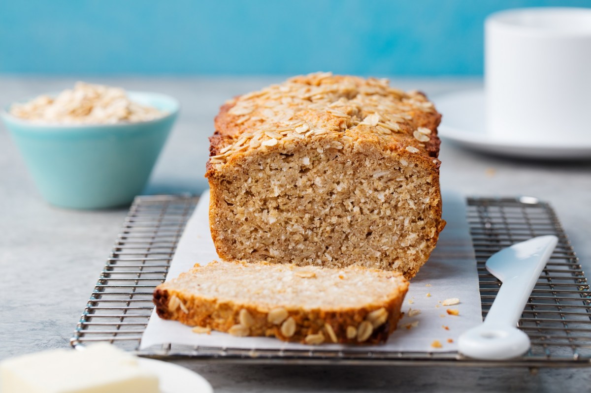 Un pan de avena cortado en una tabla, junto a él un cuchillo blanco, al fondo desenfocado un tazón con hojuelas de avena.