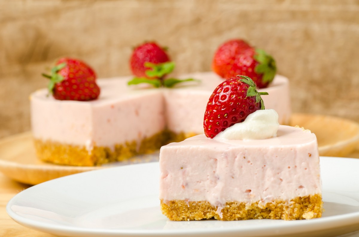 Un trozo de pastel de mousse de fresa, decorado con una fresa, en un plato, con el pastel cortado al fondo.