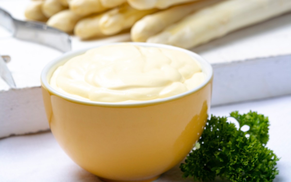 Un tazón amarillo con salsa holandesa. Al fondo se ven espárragos blancos.