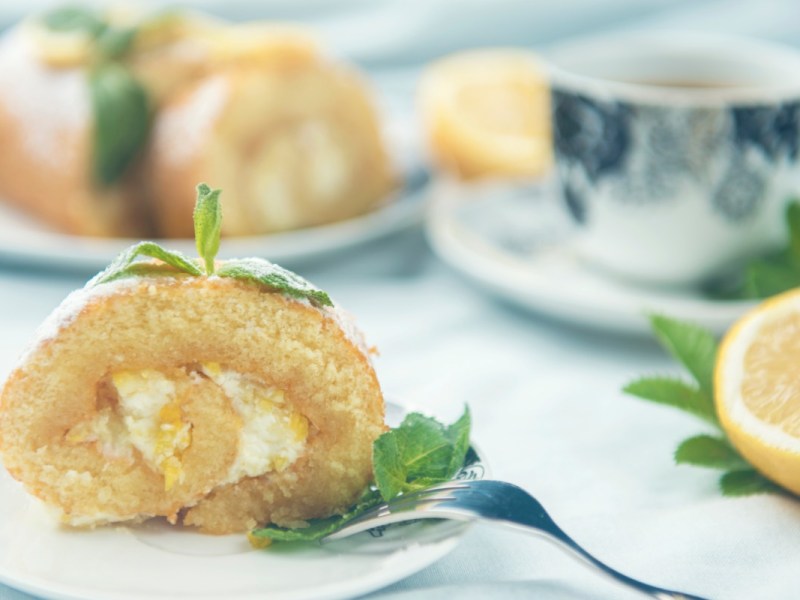 Una rebanada de brazo de gitano de limón, decorada con menta, en un plato. Al fondo se ve una taza de café y el resto del pastel.