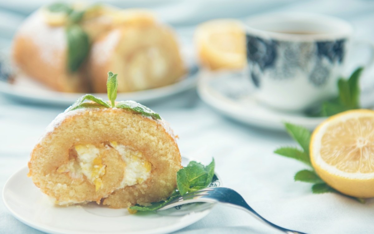 Una rebanada de brazo de gitano de limón, decorada con menta, en un plato. Al fondo se ve una taza de café y el resto del pastel.