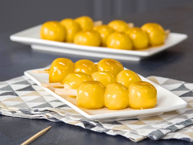 Tres brochetas de dango se encuentran en un plato blanco. Al fondo se ven más brochetas de dango. Están cubiertas con un glaseado dorado.