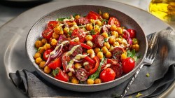 Un plato hondo grande con ensalada de garbanzos tostados con chorizo. El plato está sobre un plato más grande. También hay un tenedor sobre una servilleta de tela.