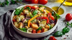Un plato grande de ensalada de pan con pimientos asados. Alrededor se ven ingredientes frescos como albahaca y tomates. Además, hay un tazón con aceite y una cuchara en la encimera.