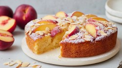 Una tabla redonda blanca sobre la que se coloca un pastel redondo de nectarina y suero de leche cortado. El pastel está espolvoreado con azúcar glas. Al fondo se ven nectarinas. En primer plano hay algunas almendras laminadas.