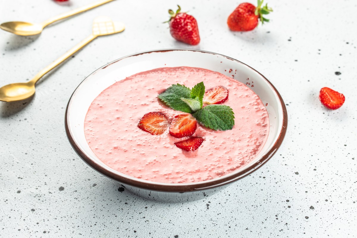 Un tazón de sopa de fresa decorado con fresas y menta, junto a unas fresas y cucharas doradas.