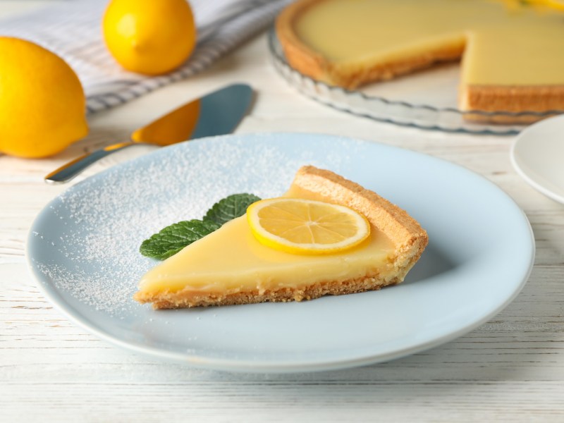 Tarta de limón, adornada con menta y una rodaja de limón, en un plato azul claro. Al fondo, borroso, el resto de la tarta.