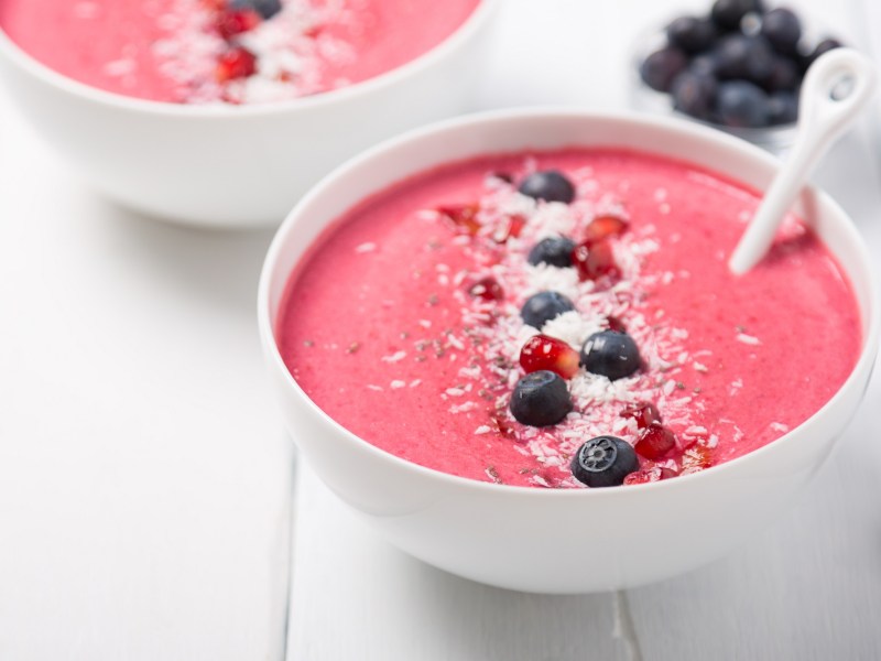 2 tazones de smoothie de fresa decorados con arándanos azules, coco rallado y semillas de granada.