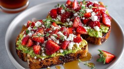 Dos tostadas de aguacate con fresas y queso fresco de cabra, decoradas con tomillo fresco. Las tostadas están en un plato gris.