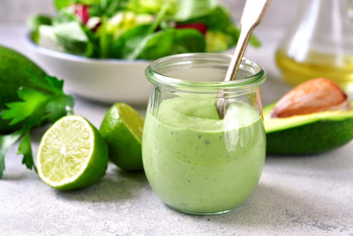 Un vaso de aderezo de aguacate y yogur con cuchara, alrededor una mitad de lima y un aguacate.