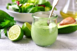 Un vaso de aderezo de aguacate y yogur con cuchara, alrededor una mitad de lima y un aguacate.