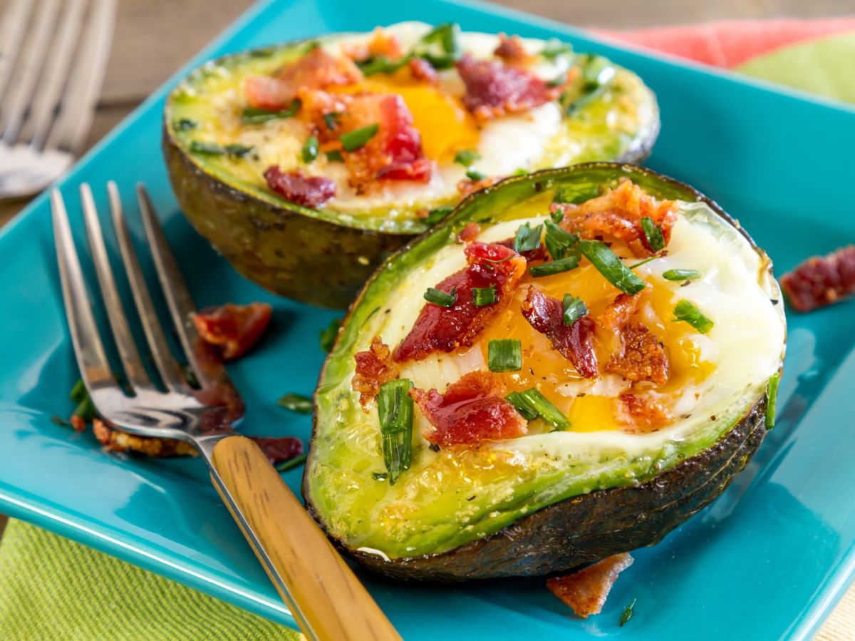 Aguacates con huevo cortados, rellenos con tocino y hierbas. Un tenedor amarillo junto al plato.
