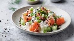 Un plato con ensalada de pepino con queso cottage y salmón. Todo ello adornado con sésamo negro, cebolletas y eneldo.