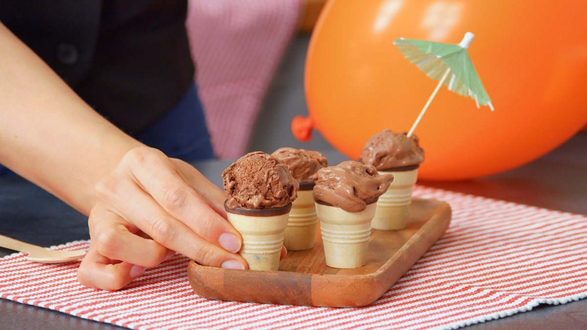 Una pequeña tabla de madera que está sobre un mantel individual a rayas rojas. En la tabla hay algunos barquillos con helado de chocolate casero. En una bola de helado hay una pequeña sombrilla de cóctel. Una mano agarra uno de los barquillos de helado. Al fondo se puede ver un globo naranja.