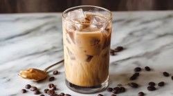 Un vaso de latte helado de mantequilla de cacahuete sobre una superficie de trabajo de mármol claro. Alrededor hay granos de café esparcidos y se ve una cuchara llena de mantequilla de cacahuete.