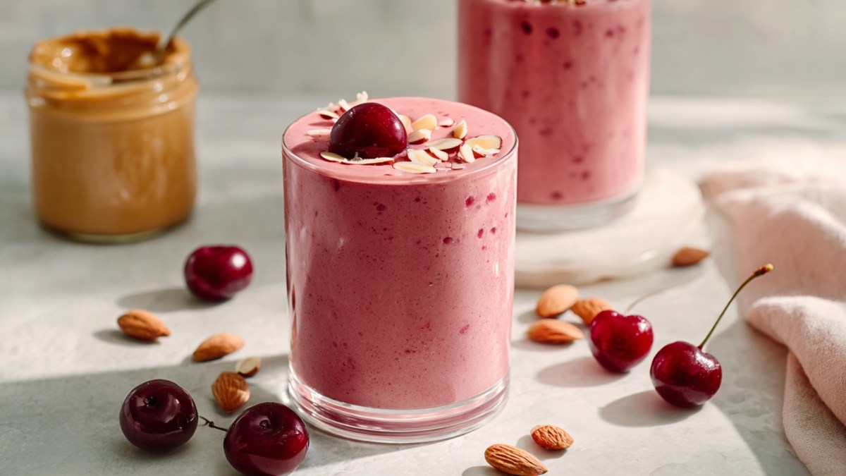Un vaso de smoothie de cereza y almendra, decorado con una cereza fresca y algunas almendras fileteadas. Alrededor hay almendras marrones y algunas cerezas esparcidas. Al fondo se puede ver otro vaso con smoothie y un vaso con crema de almendras, con una cuchara dentro.