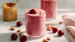 Un vaso de smoothie de cereza y almendra, decorado con una cereza fresca y algunas almendras fileteadas. Alrededor hay almendras marrones y algunas cerezas esparcidas. Al fondo se puede ver otro vaso con smoothie y un vaso con crema de almendras, con una cuchara dentro.