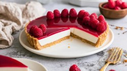 Un plato con una tarta de panna cotta de frambuesa cortada y decorada con frambuesas frescas. Junto al plato hay un viejo tenedor para pastel. Al fondo se ve un bol con frambuesas. En primer plano se puede ver un trozo de tarta en un plato.