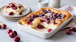 Un molde para hornear con una tarta de queso y cerezas con hojaldre, espolvoreada con azúcar glas. También se ve una pila de platos. En el plato superior hay un trozo de tarta. Alrededor hay cerezas frescas esparcidas, al fondo hay un vaso de leche. El molde para hornear está sobre un paño de cocina.